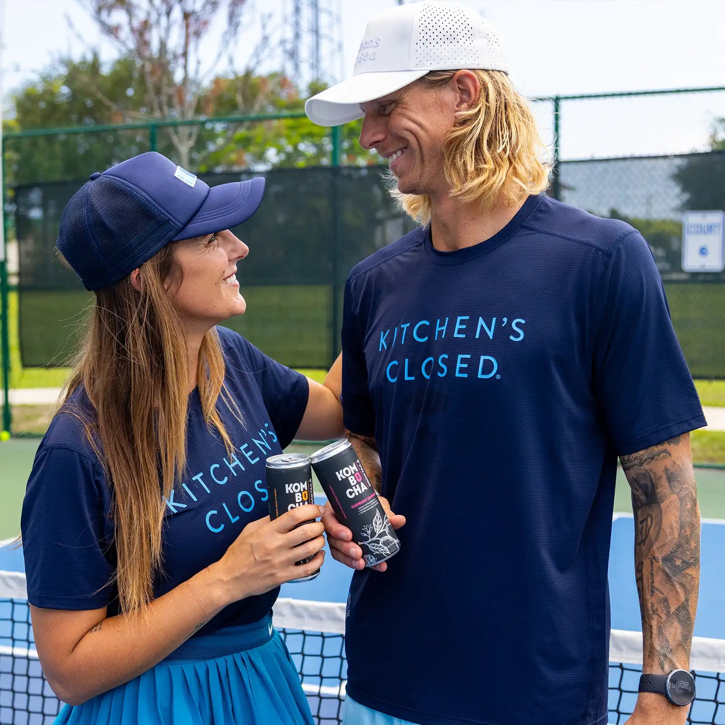 couple holding cocktails on pickleball court wearing swinton hats and navy blue luxe kitchen's closed tees