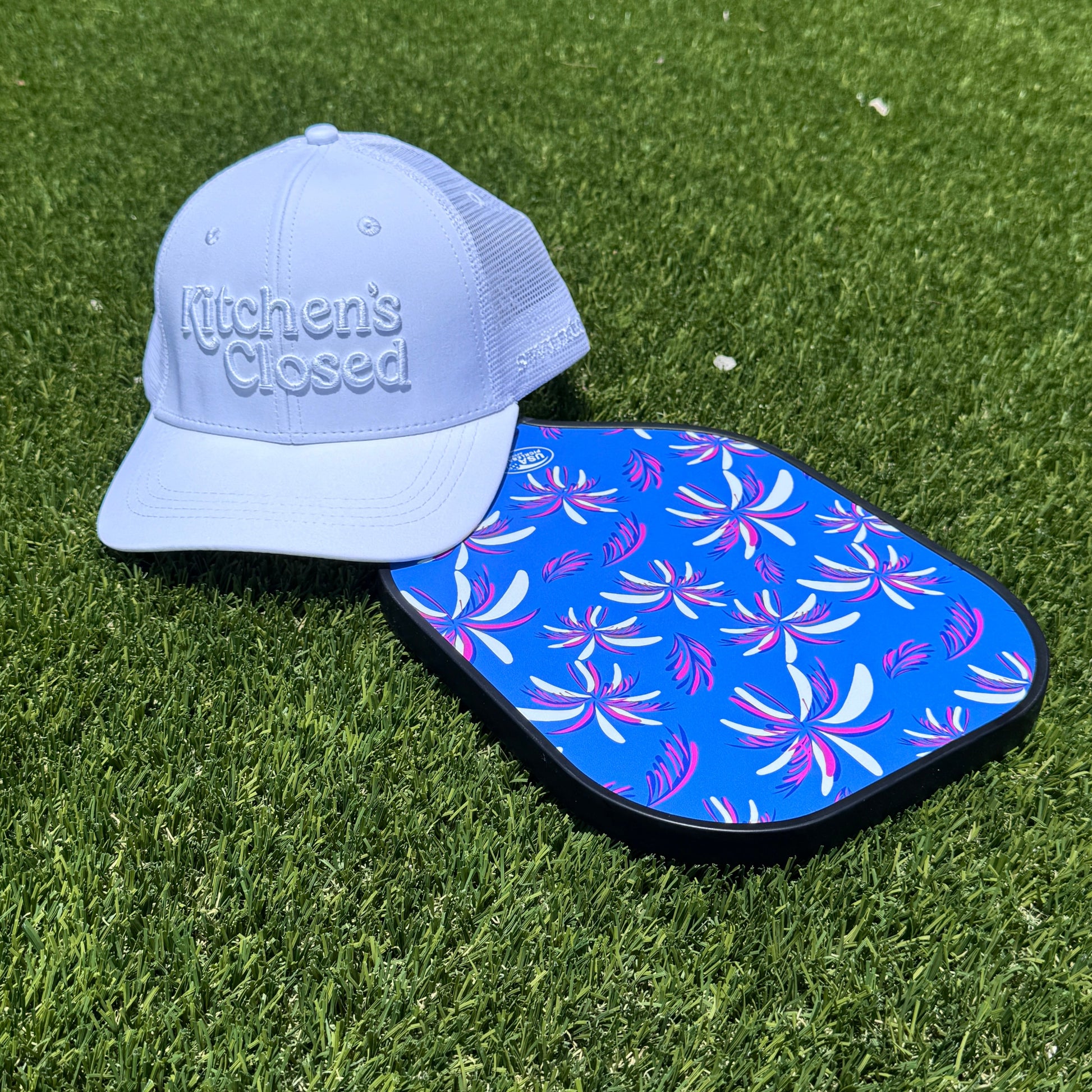 white pickleball hat laying on blue paddle on turf